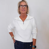 Woman wearing a white sweater and dark pants against a plain background