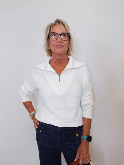 Woman wearing a white sweater and dark pants against a plain background