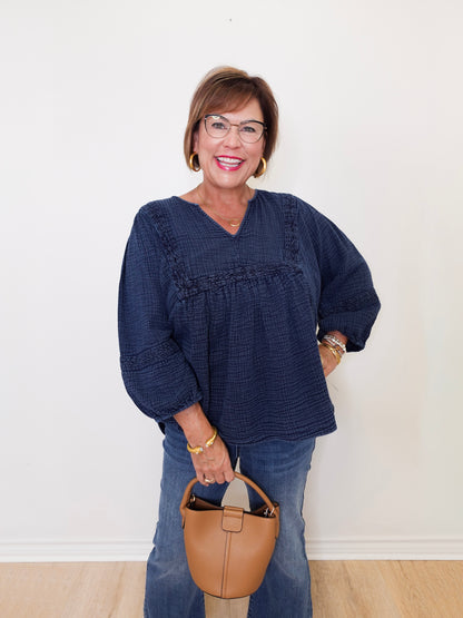 Woman wearing a blue top and jeans, holding a tan handbag against a white background