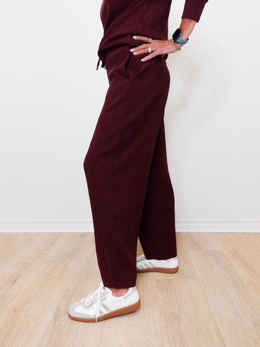 Person wearing burgundy pants and white sneakers on a light wooden floor.