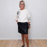 Woman wearing a white sweater with black floral designs and a black skirt against a white wall.