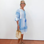Woman wearing a blue and white striped dress holding a beige handbag against a white wall.