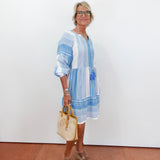 Woman wearing a blue and white striped dress holding a beige handbag against a white wall.