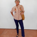 Woman wearing a beige embroidered blouse, blue pants, and black sandals against a white wall.