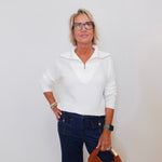 Woman wearing a white sweater and dark jeans, holding a brown handbag against a white background
