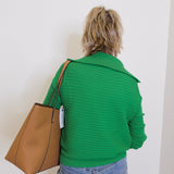 Person wearing a green sweater holding a brown bag against a white wall