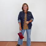A woman stands against a plain white wall wearing a dark blue denim jacket over a gold and green patterned blouse, light blue flared jeans, and dark sandals. She is holding a small dark red handbag.