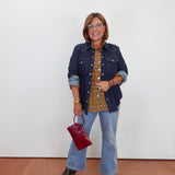 A woman stands against a plain white wall wearing a dark blue denim jacket over a gold and green patterned blouse, light blue flared jeans, and dark sandals. She is holding a small dark red handbag.