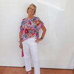 Woman wearing a colorful floral top and white pants against a white wall.