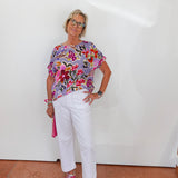 Woman wearing a colorful floral top and white pants against a white wall.