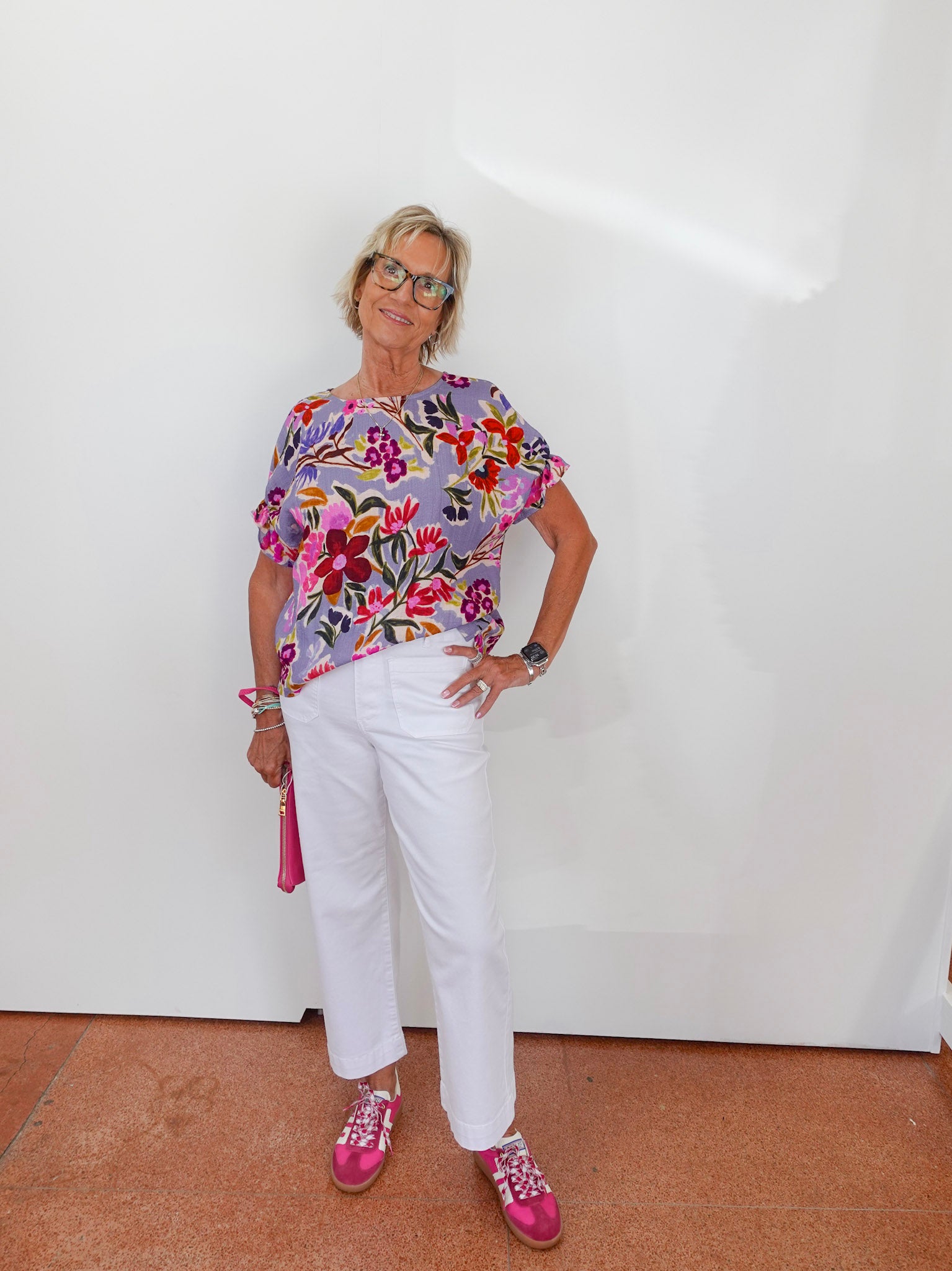 Woman wearing a colorful floral top and white pants against a white wall.