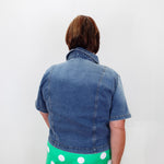 A person viewed from behind wearing a short-sleeve blue denim jacket and bright green pants with white polka dots, standing against a plain white wall.