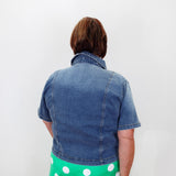 A person viewed from behind wearing a short-sleeve blue denim jacket and bright green pants with white polka dots, standing against a plain white wall.