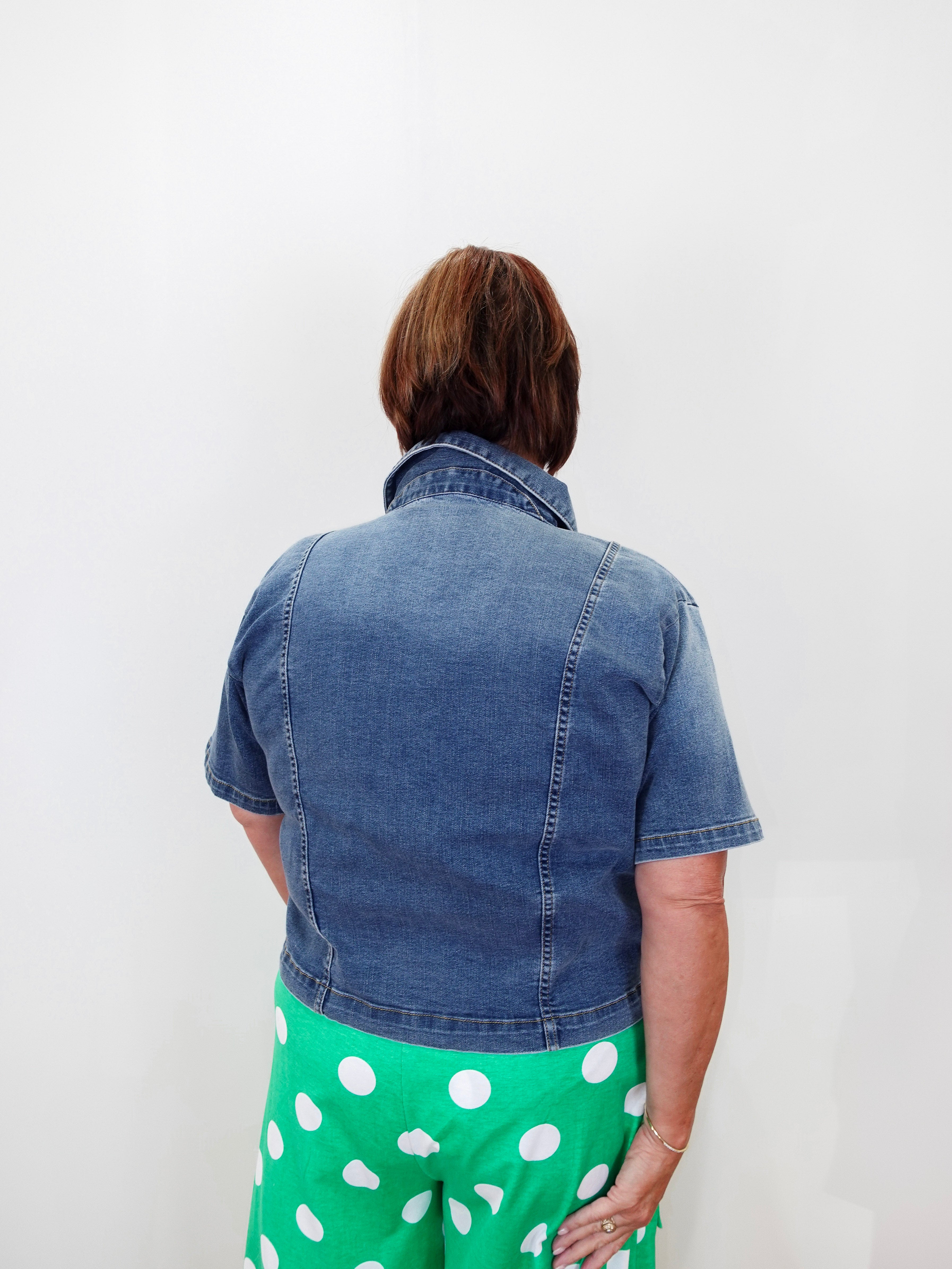 A person viewed from behind wearing a short-sleeve blue denim jacket and bright green pants with white polka dots, standing against a plain white wall.