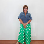 A woman stands against a plain white wall wearing a short-sleeve blue denim shirt and bright green wide-leg pants with large white polka dots. She is wearing light-colored flats and standing on a terracotta-colored tiled floor.