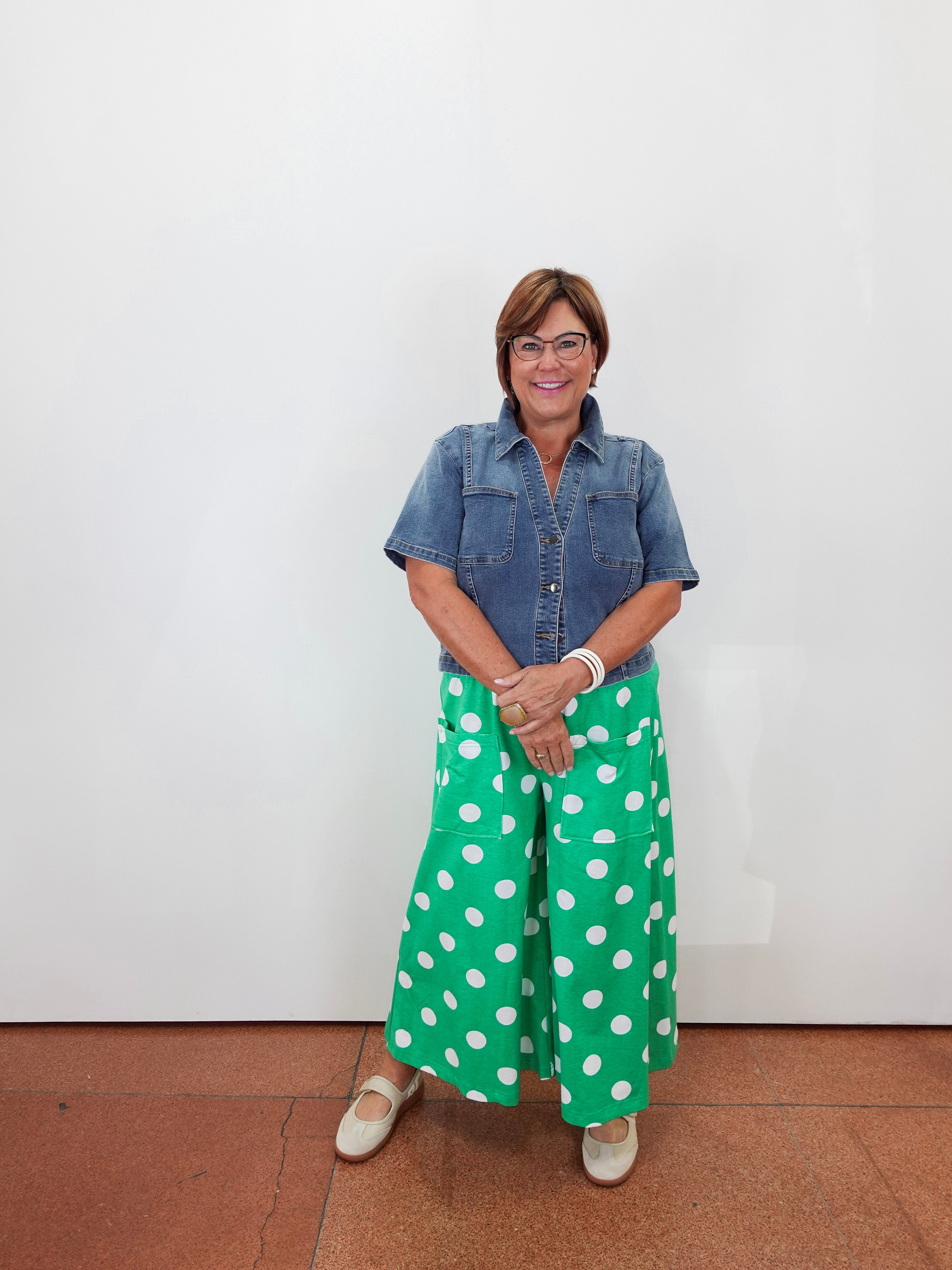 A woman stands against a plain white wall wearing a short-sleeve blue denim shirt and bright green wide-leg pants with large white polka dots. She is wearing light-colored flats and standing on a terracotta-colored tiled floor.