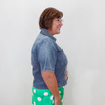 A woman shown in profile wearing a short-sleeve blue denim jacket and a bright green skirt with white polka dots, standing against a plain white wall.