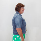 A woman shown in profile wearing a short-sleeve blue denim jacket and a bright green skirt with white polka dots, standing against a plain white wall.