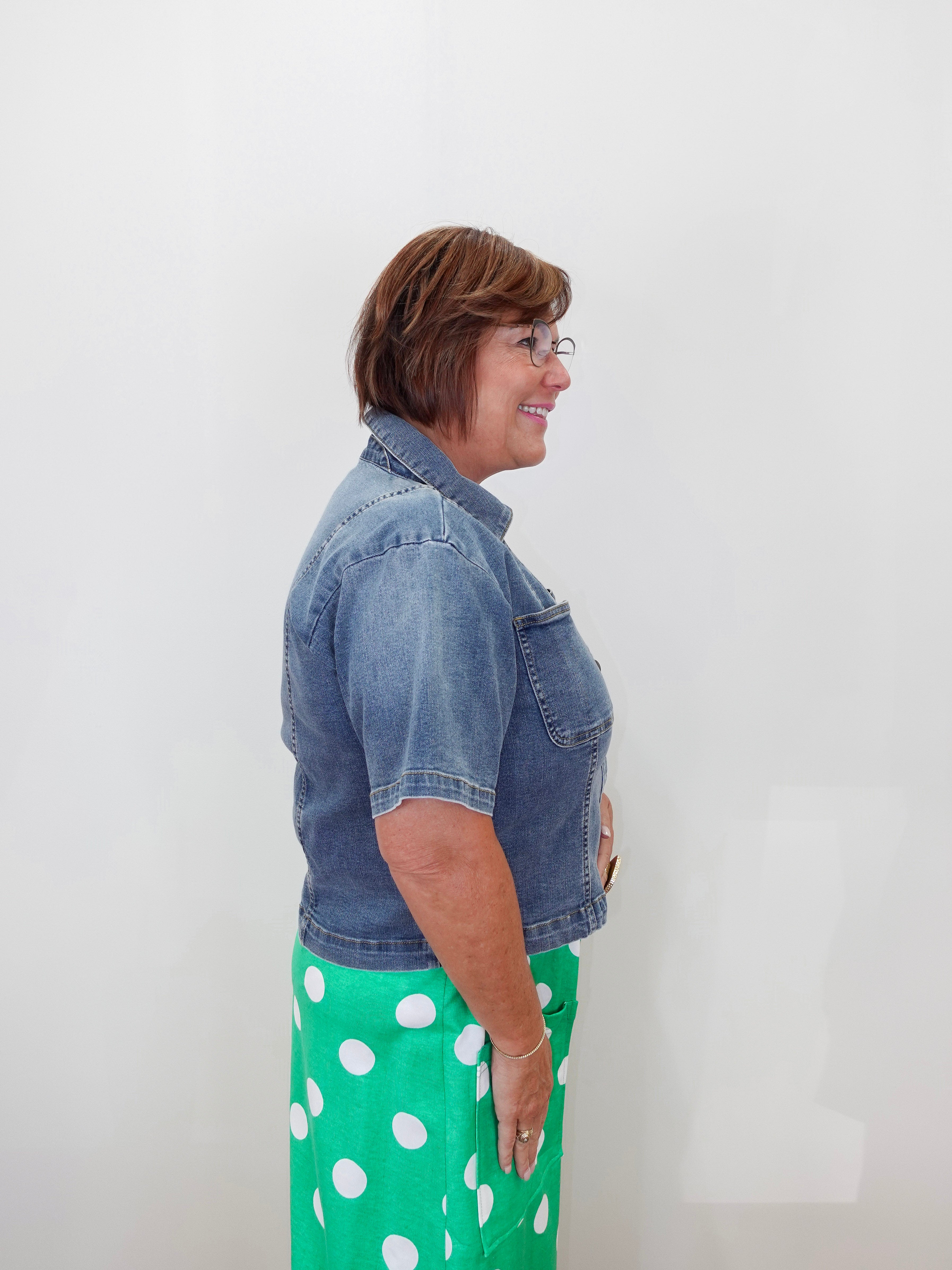 A woman shown in profile wearing a short-sleeve blue denim jacket and a bright green skirt with white polka dots, standing against a plain white wall.