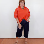 Woman wearing an orange blouse and black pants against a white wall.