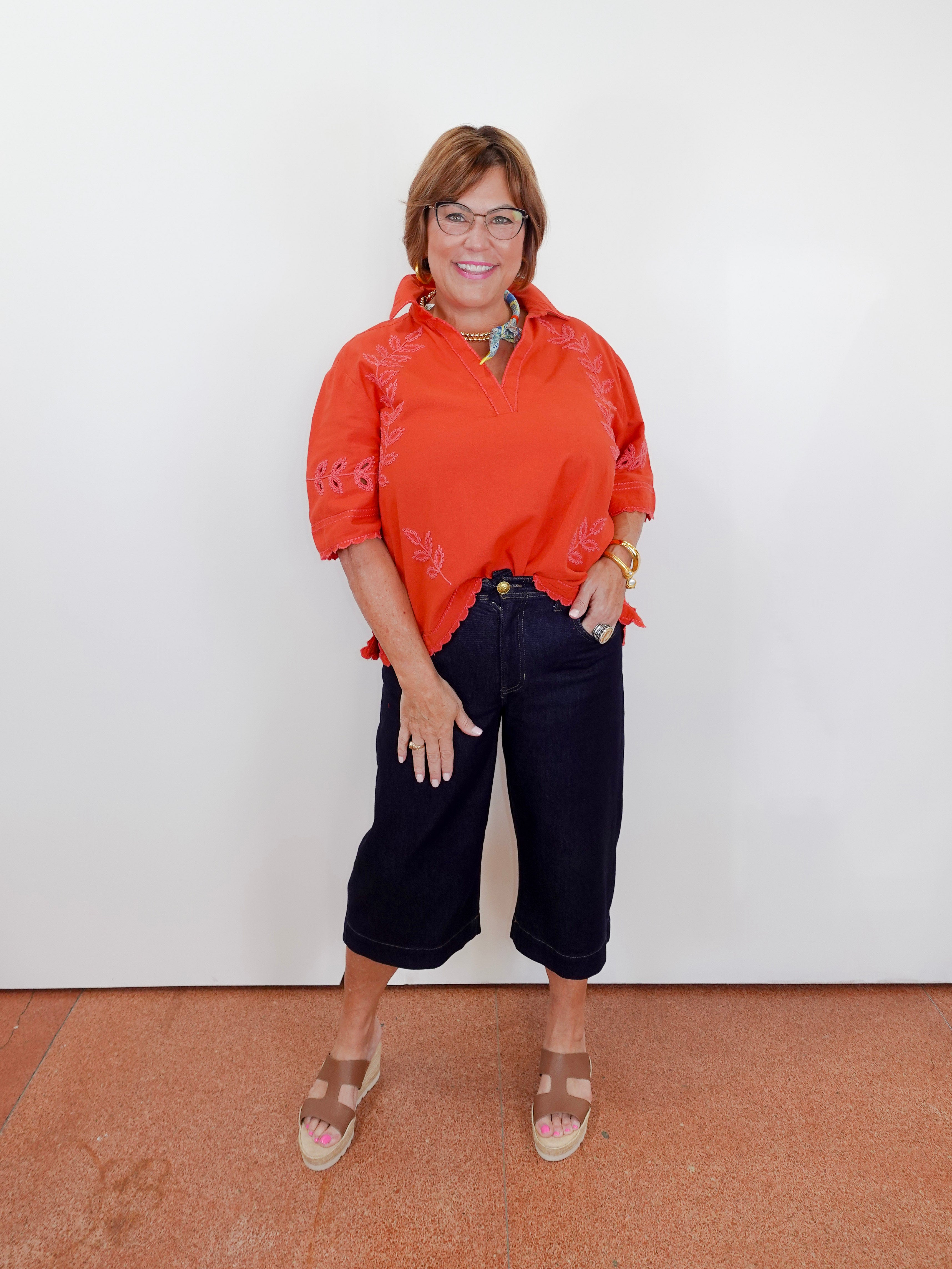 Woman wearing an orange blouse and black pants against a white wall.