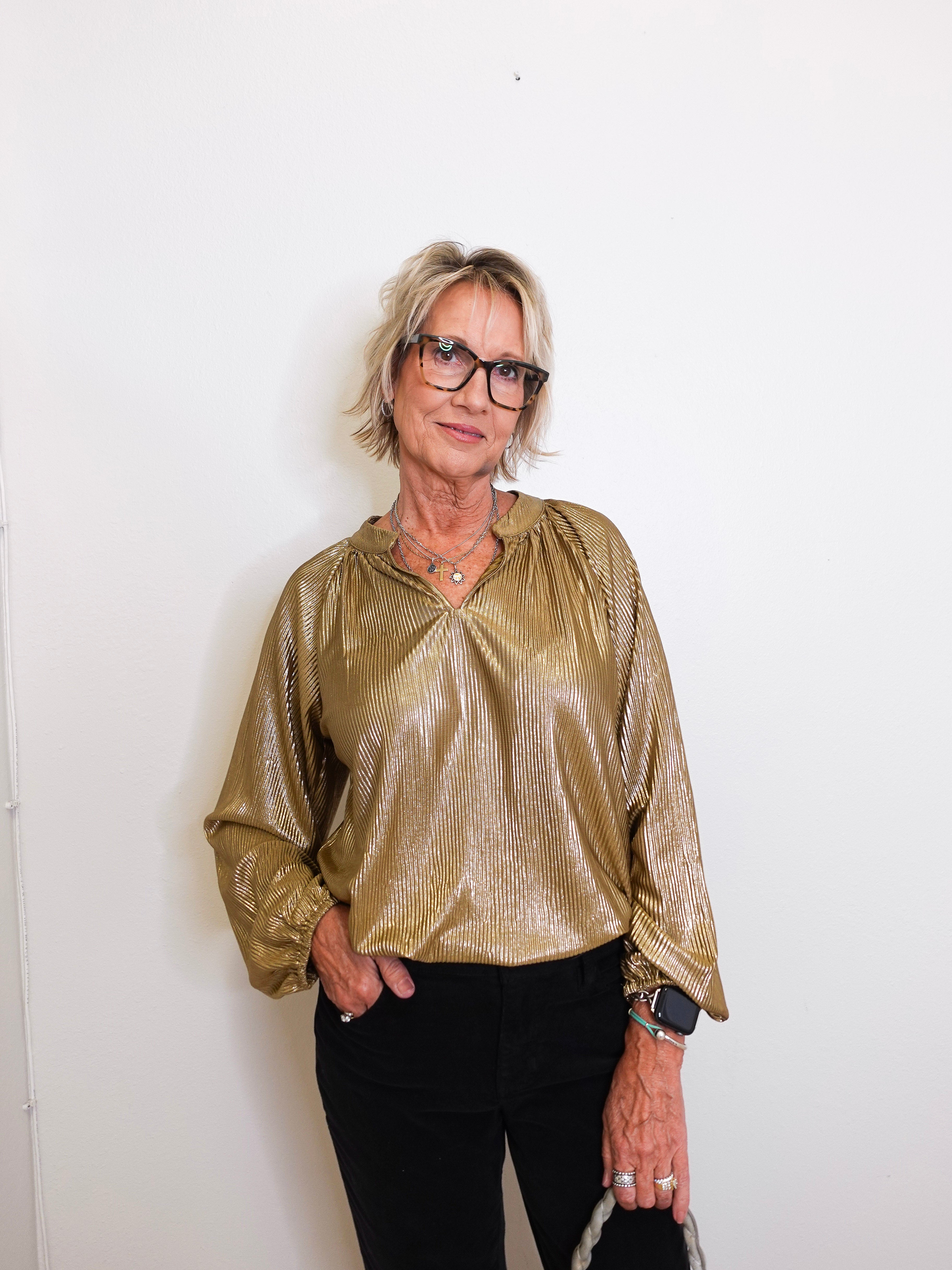 Woman wearing a gold metallic blouse and black pants against a white background