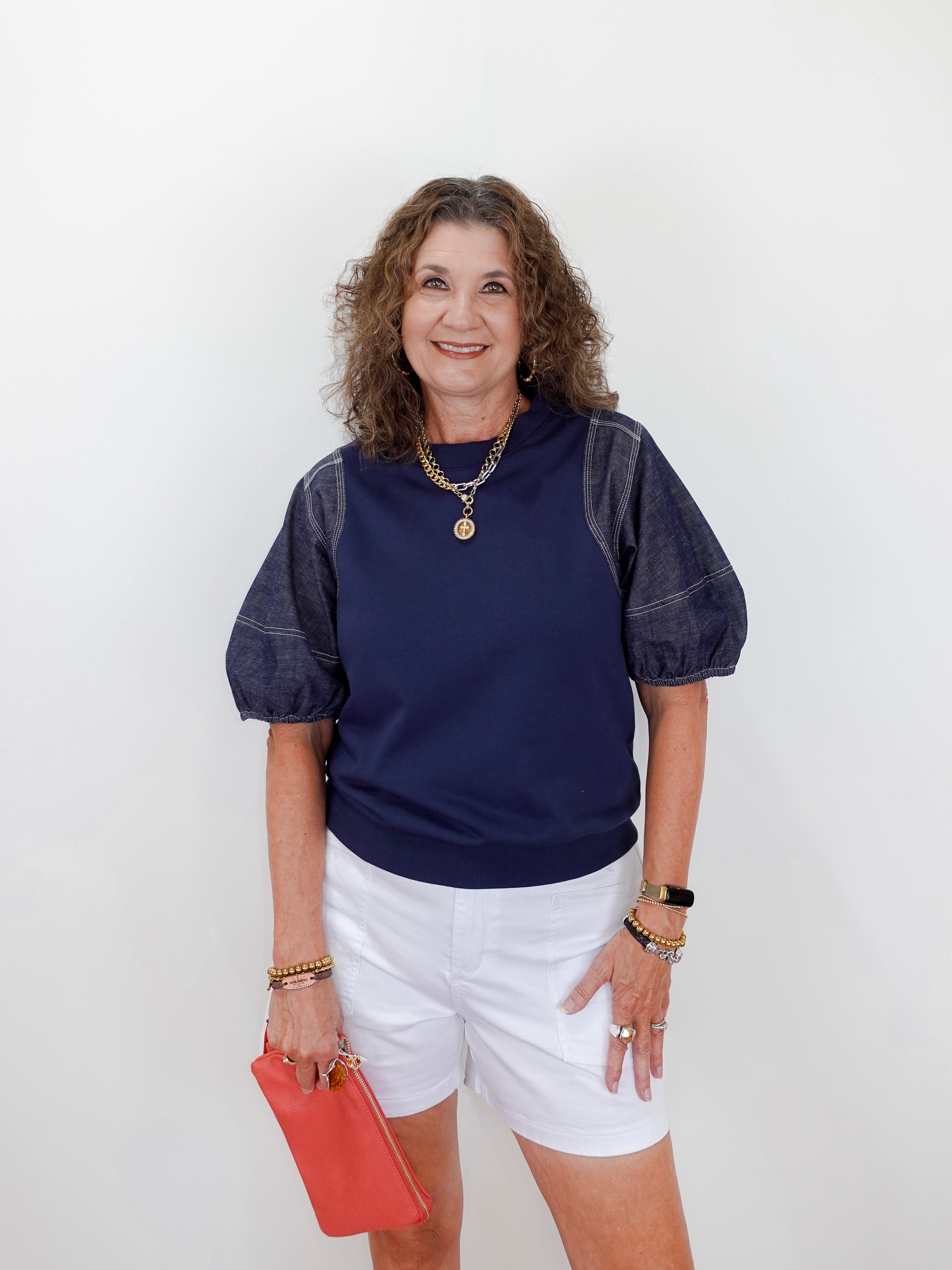 Woman wearing a navy blue top with white shorts and holding a red clutch against a white background