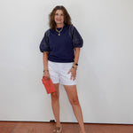 Woman wearing a navy shirt, white shorts, and sandals, holding a red clutch against a white wall.