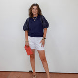 Woman wearing a navy shirt, white shorts, and sandals, holding a red clutch against a white wall.