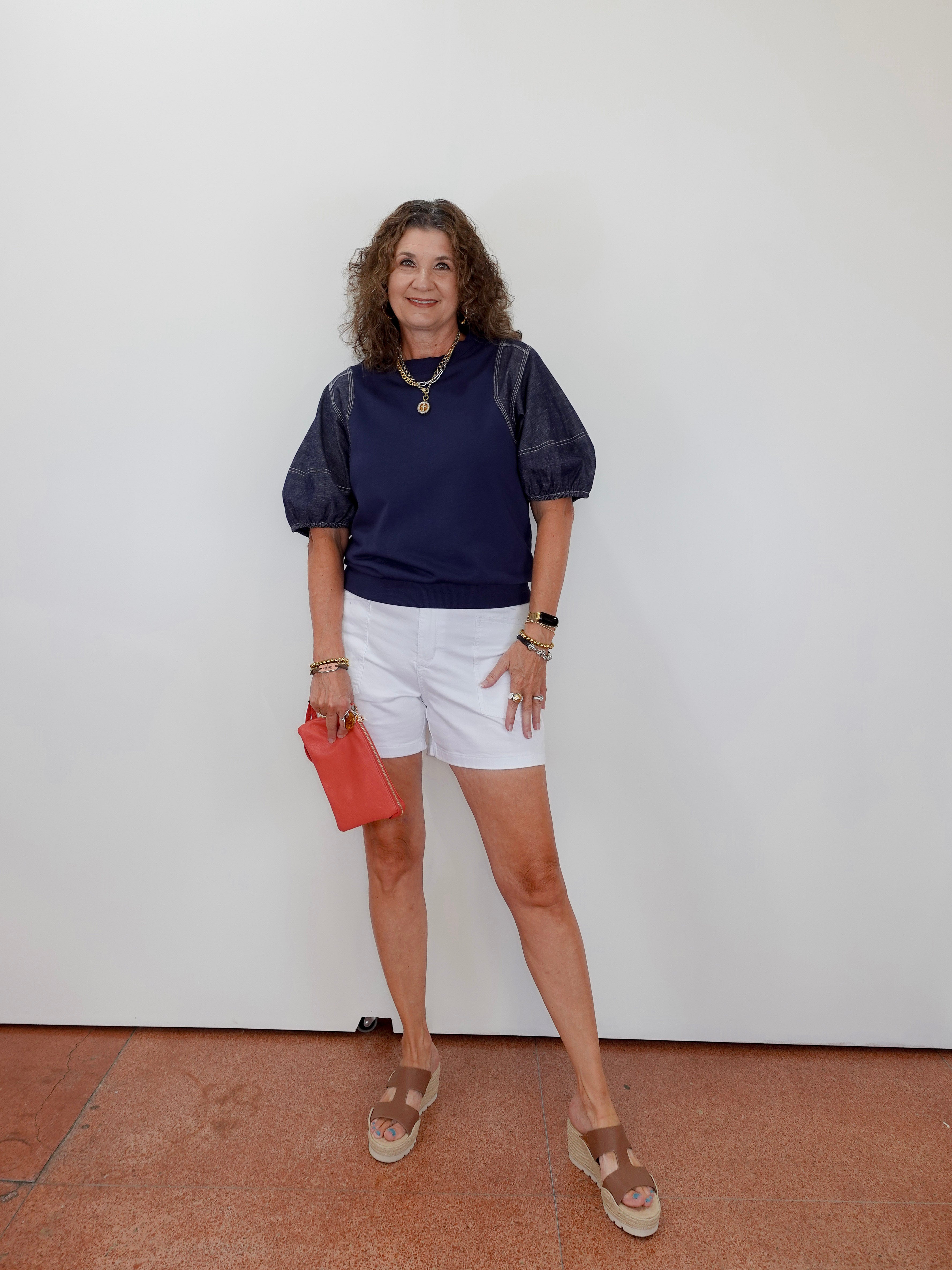 Woman wearing a navy shirt, white shorts, and sandals, holding a red clutch against a white wall.