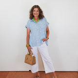 Woman wearing a light blue polka dot shirt, white pants, and brown sandals, holding a beige handbag against a white wall.