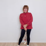 Woman wearing a red sweater and black leggings standing against a white wall.