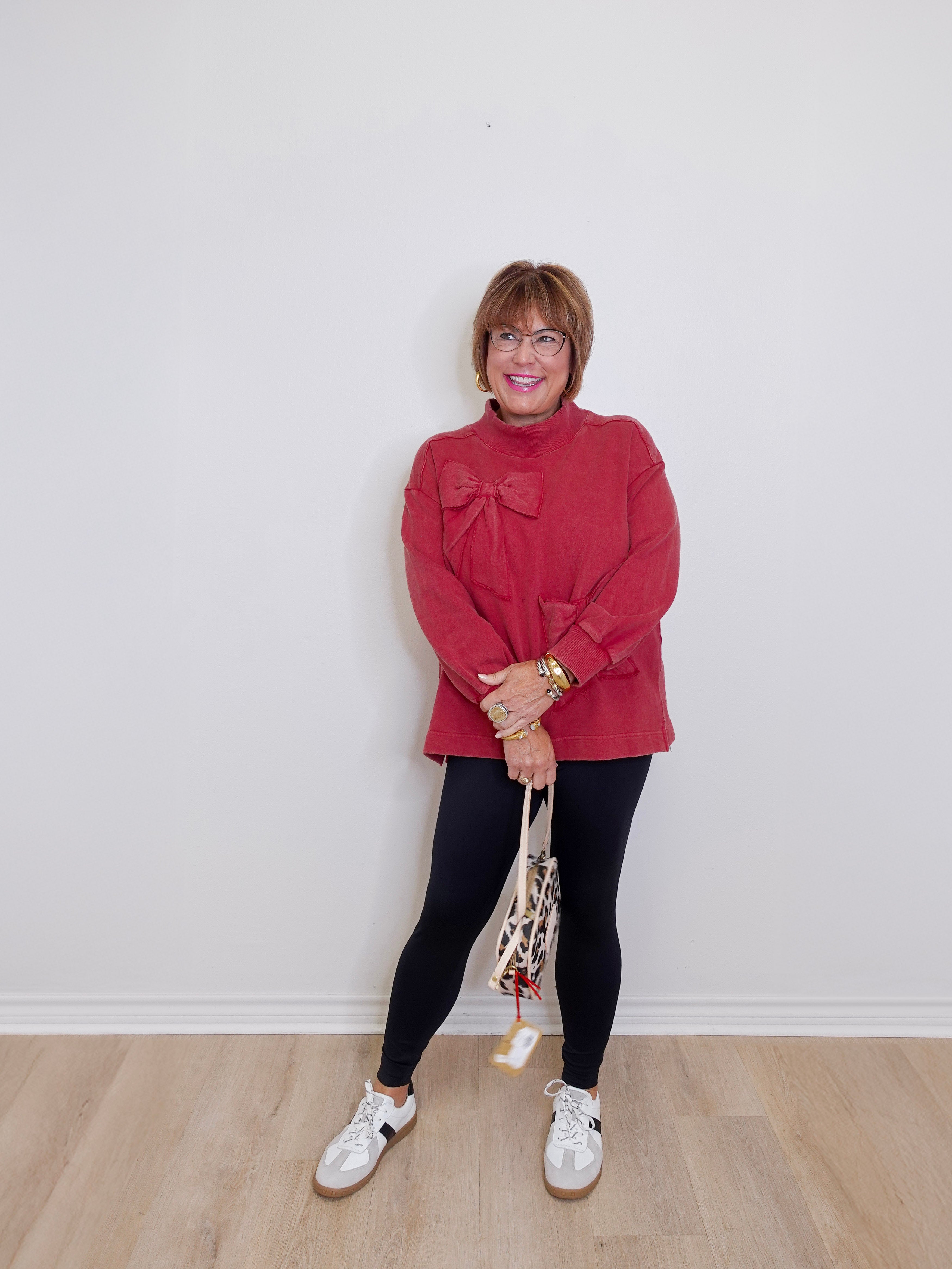 Woman wearing a red sweater and black leggings standing against a white wall.