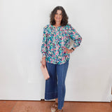 Woman wearing a floral blouse and blue jeans standing against a white wall.