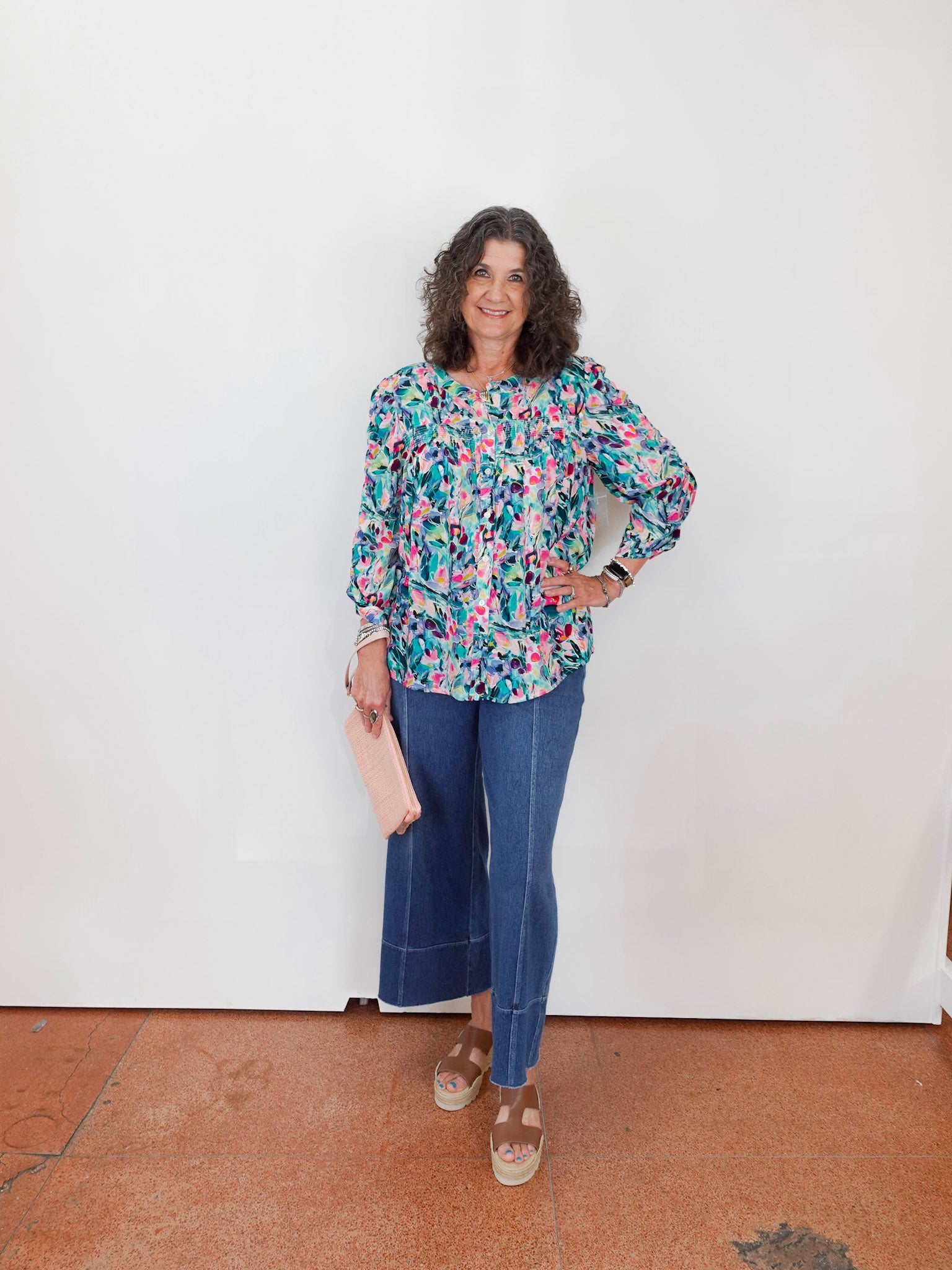 Woman wearing a floral blouse and blue jeans standing against a white wall.