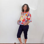 Woman wearing a colorful floral blouse and navy shorts standing against a white wall.