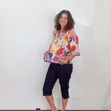 Woman wearing a colorful floral blouse and navy shorts standing against a white wall.