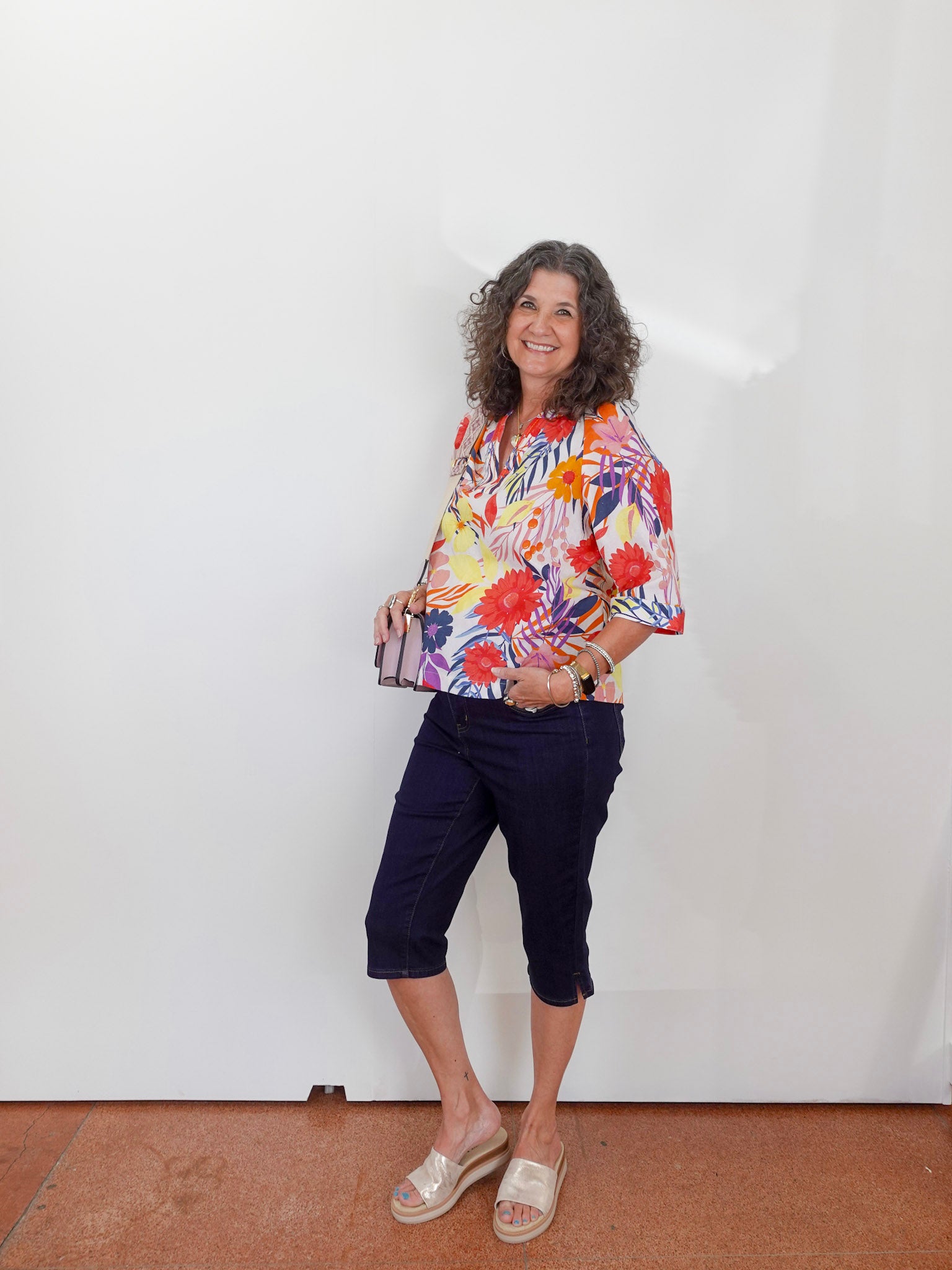 Woman wearing a colorful floral blouse and navy shorts standing against a white wall.