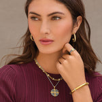 Woman wearing gold jewelry including earrings, necklace, ring, and bracelet against a neutral background