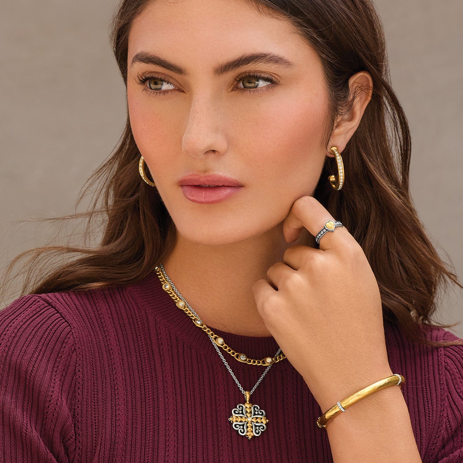 Woman wearing gold jewelry including earrings, necklace, ring, and bracelet against a neutral background