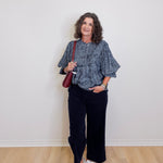 Woman wearing a blue patterned top and black pants, holding a red bag against a white wall.