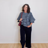 Woman wearing a blue patterned top and black pants, holding a red bag against a white wall.