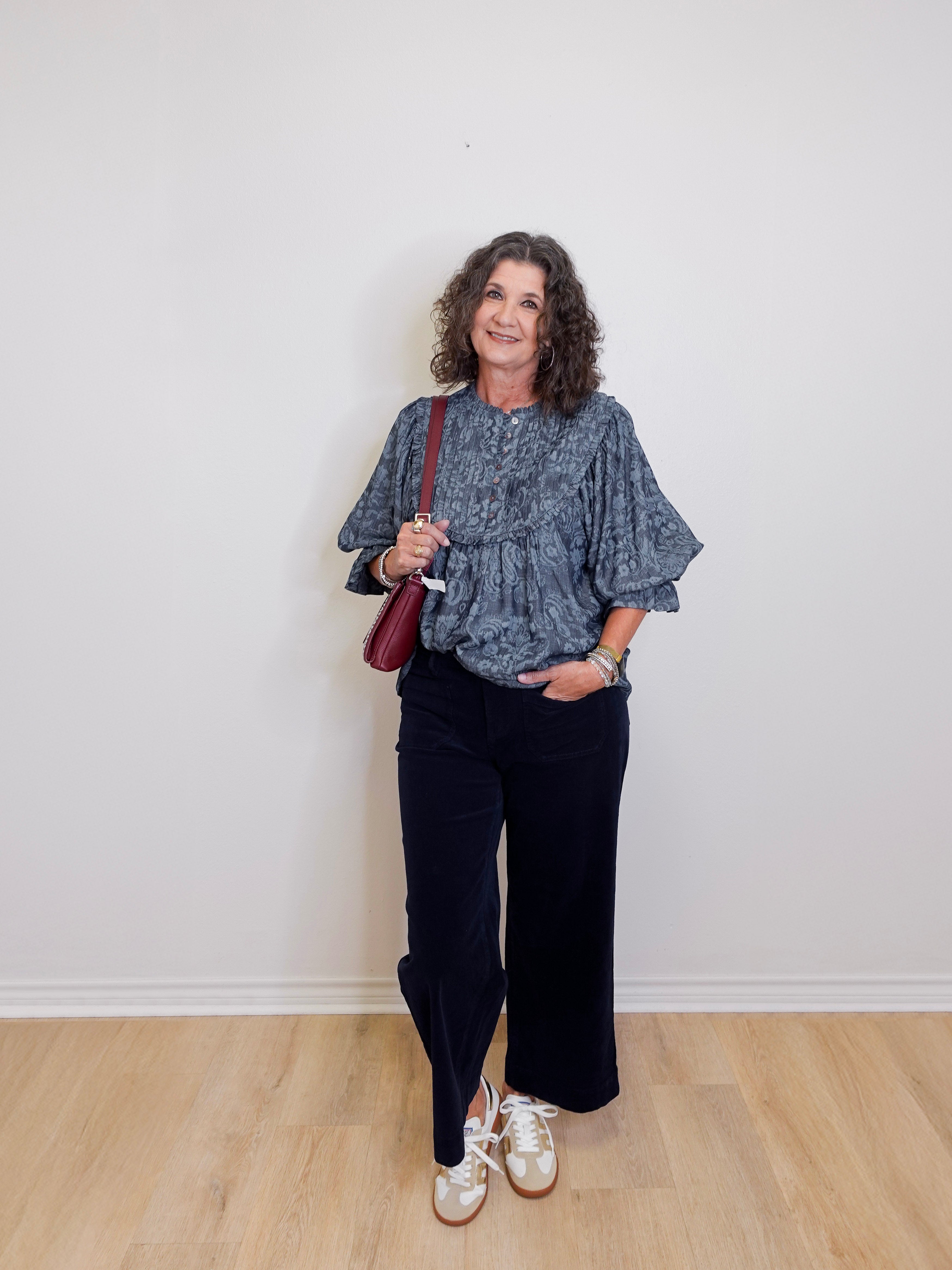 Woman wearing a blue patterned top and black pants, holding a red bag against a white wall.