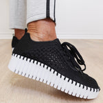 Black knitted sneakers with white soles worn by a person on a light wooden floor.