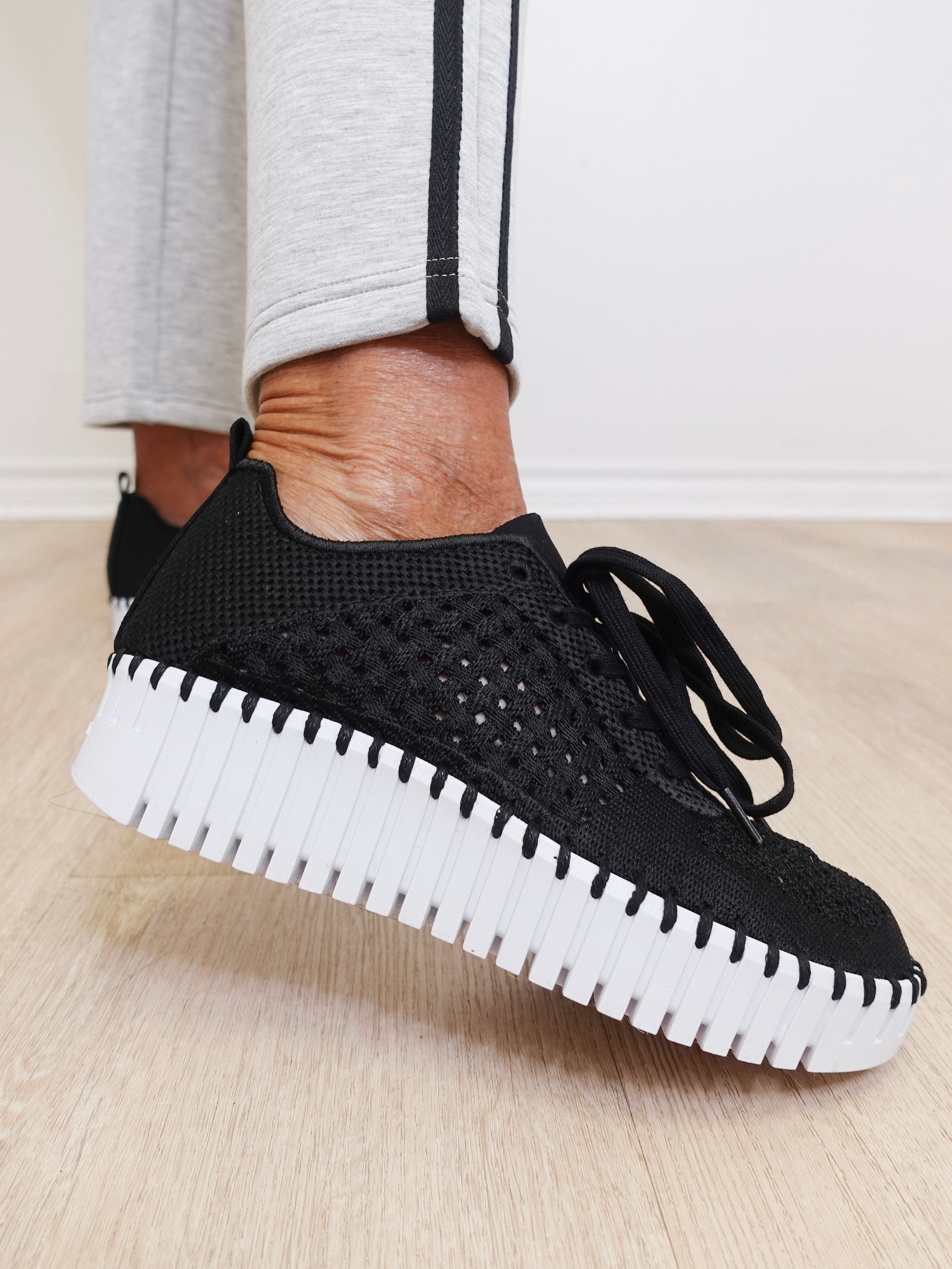 Black knitted sneakers with white soles worn by a person on a light wooden floor.