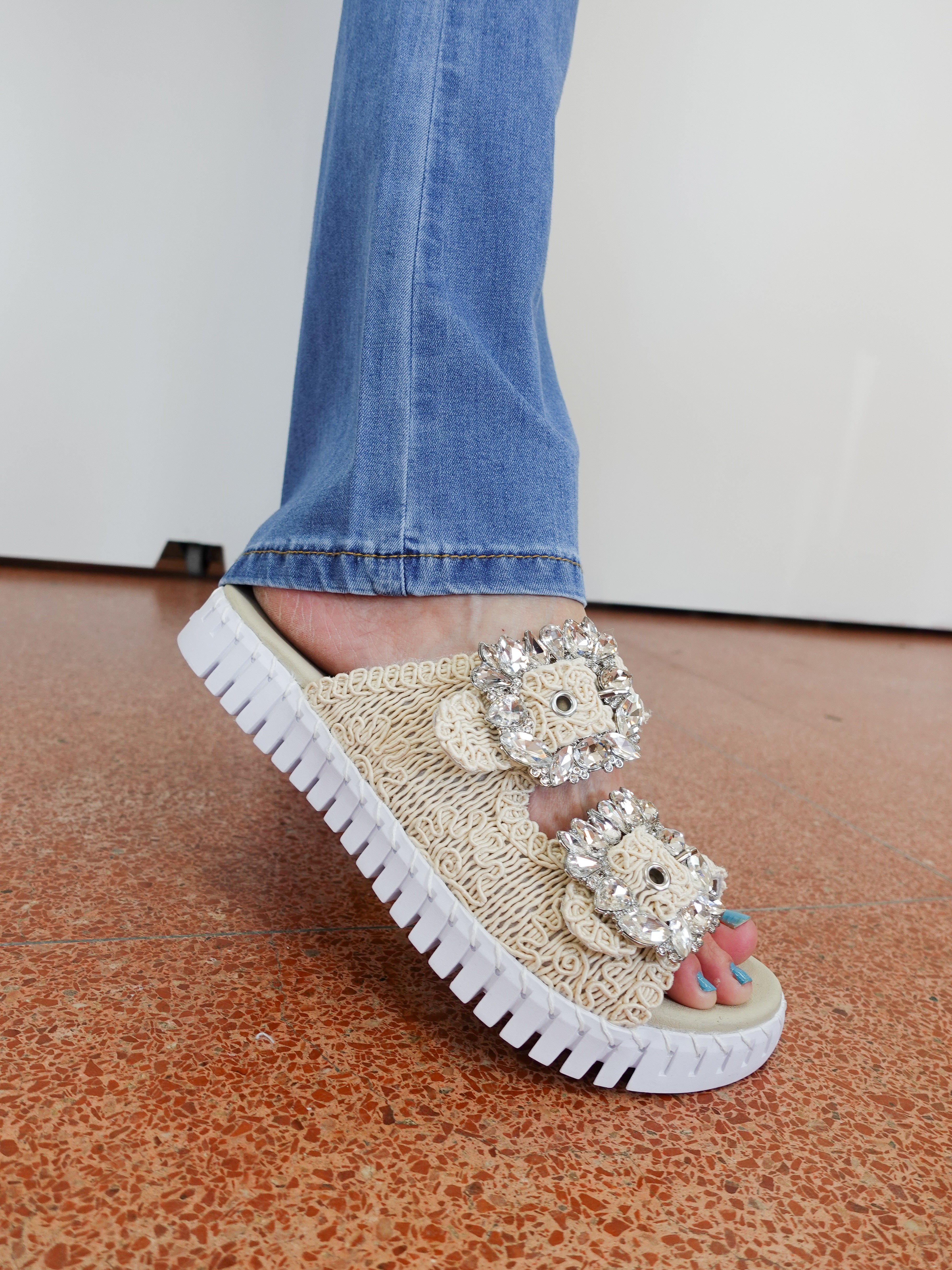 Beige sandal with rhinestone embellishments worn by a person in blue jeans on a brown floor.