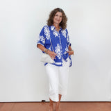 Woman wearing a blue patterned top and white pants against a white wall.
