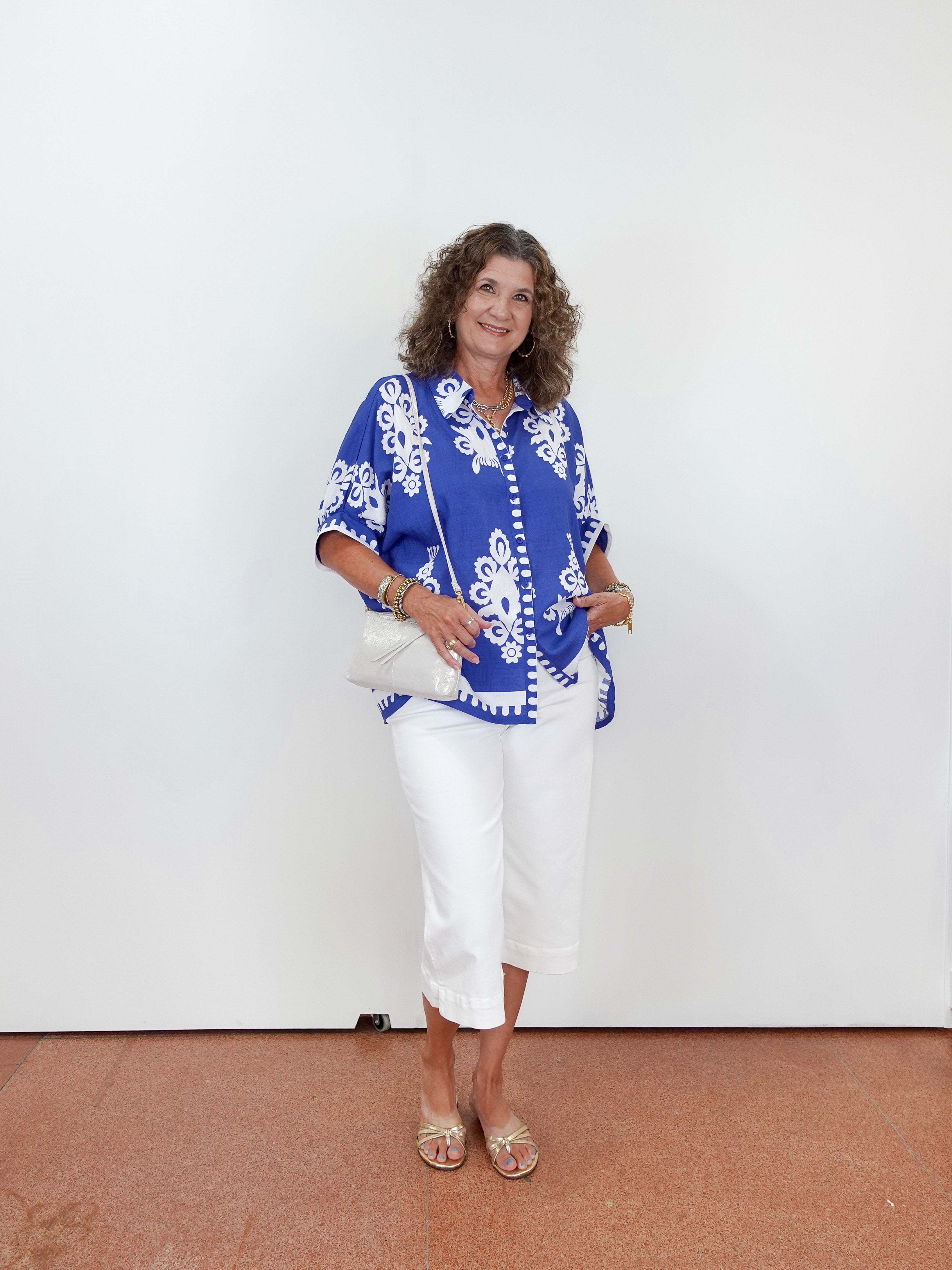 Woman wearing a blue patterned top and white pants against a white wall.