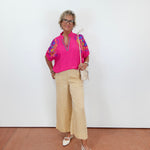 Woman wearing a bright pink top with colorful patterns and beige pants against a white wall.