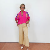 Woman wearing a bright pink top with colorful patterns and beige pants against a white wall.
